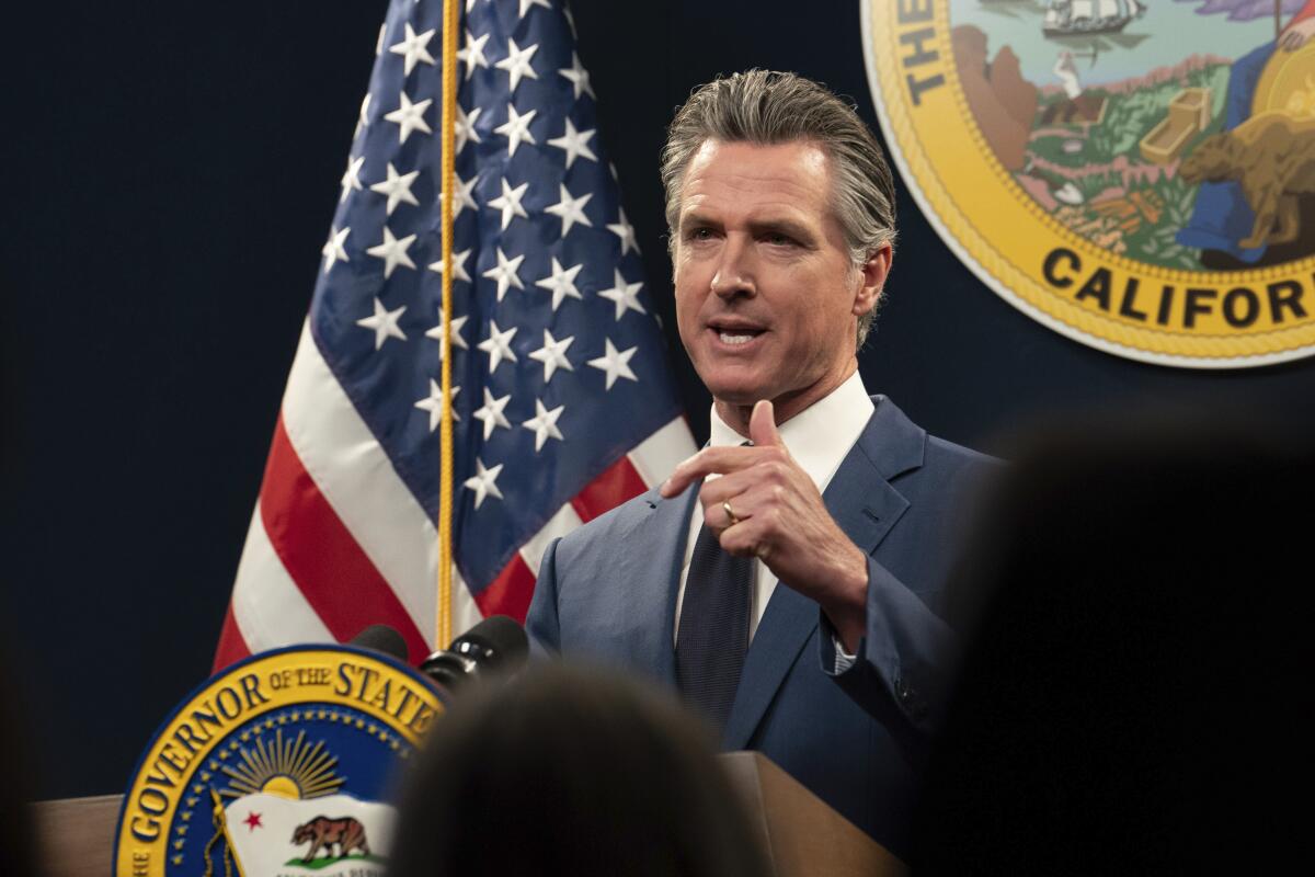 California Gov. Gavin Newsom, in a dark blue suit, speaks at a lectern with a state insignia and the American flag behind him
