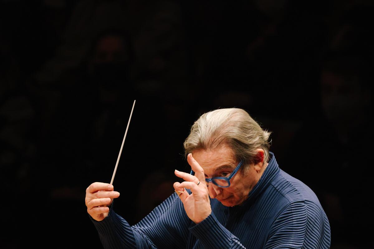A man wearing a blue turtleneck sweater and blue eyeglasses conducts with a baton.