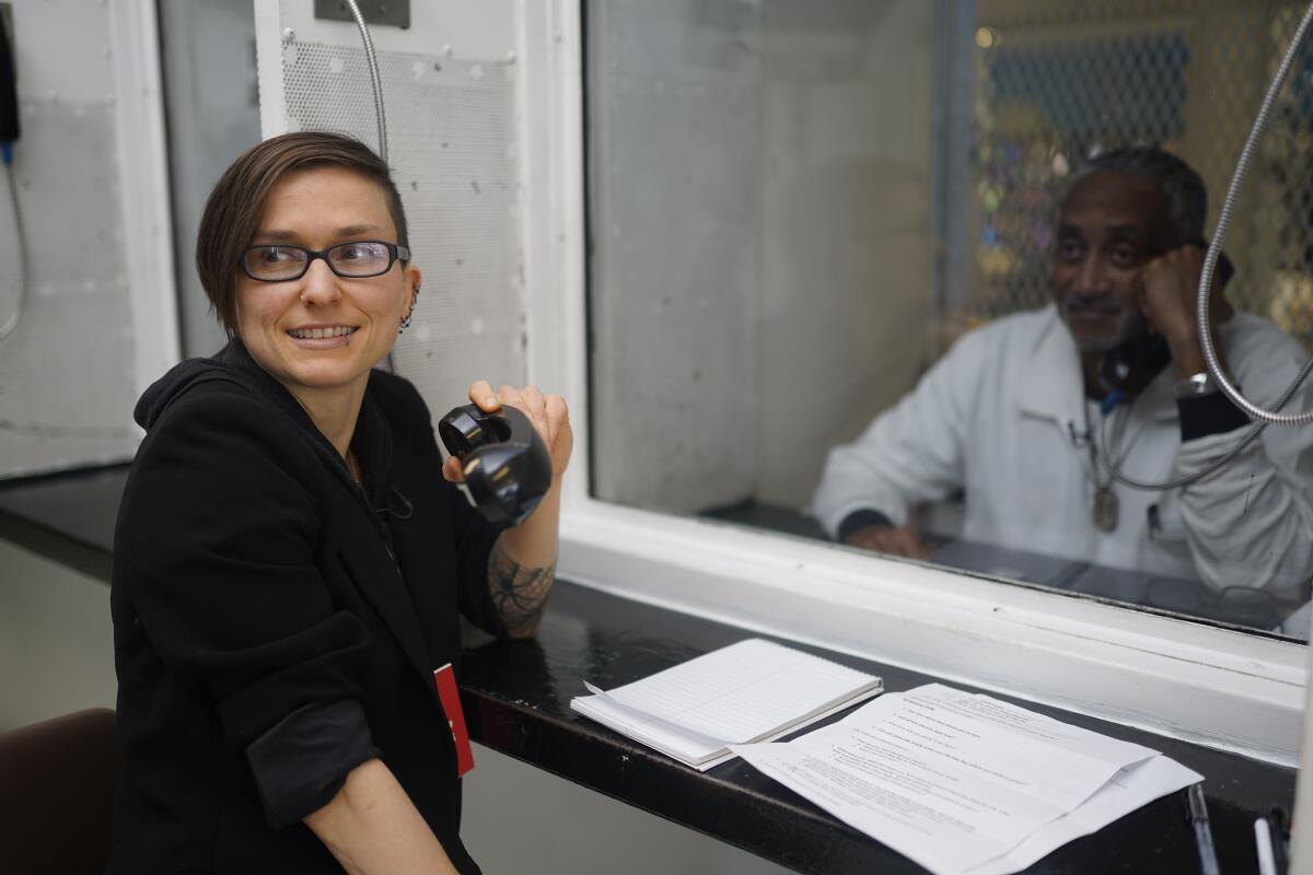 A woman with a camera in a prison, talking to an inmate through glass.