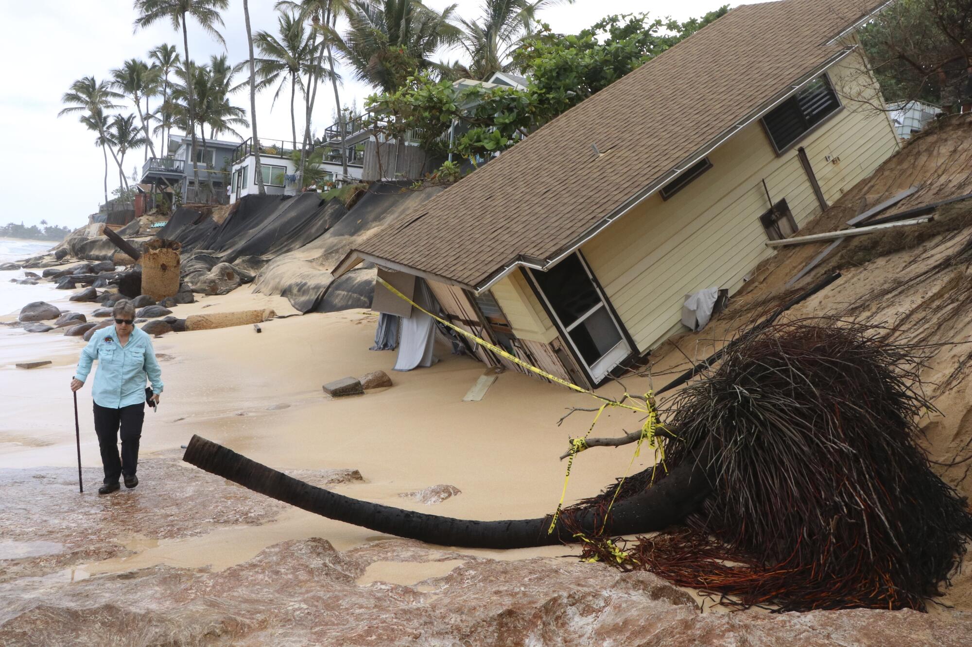 A person with a walking stick passes a home collapsed onto a beach while palm trees stand nearby and one lies on the sand.