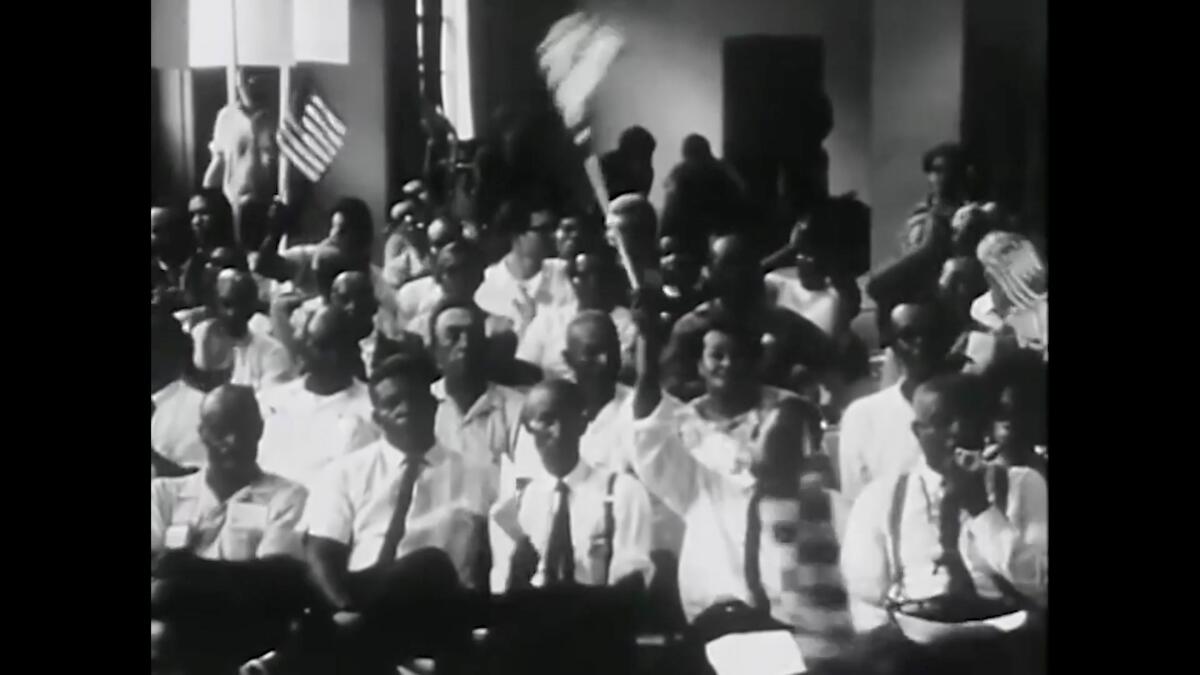 A black-and-white image from video shows a group of Black people in white shirts people waving flags.