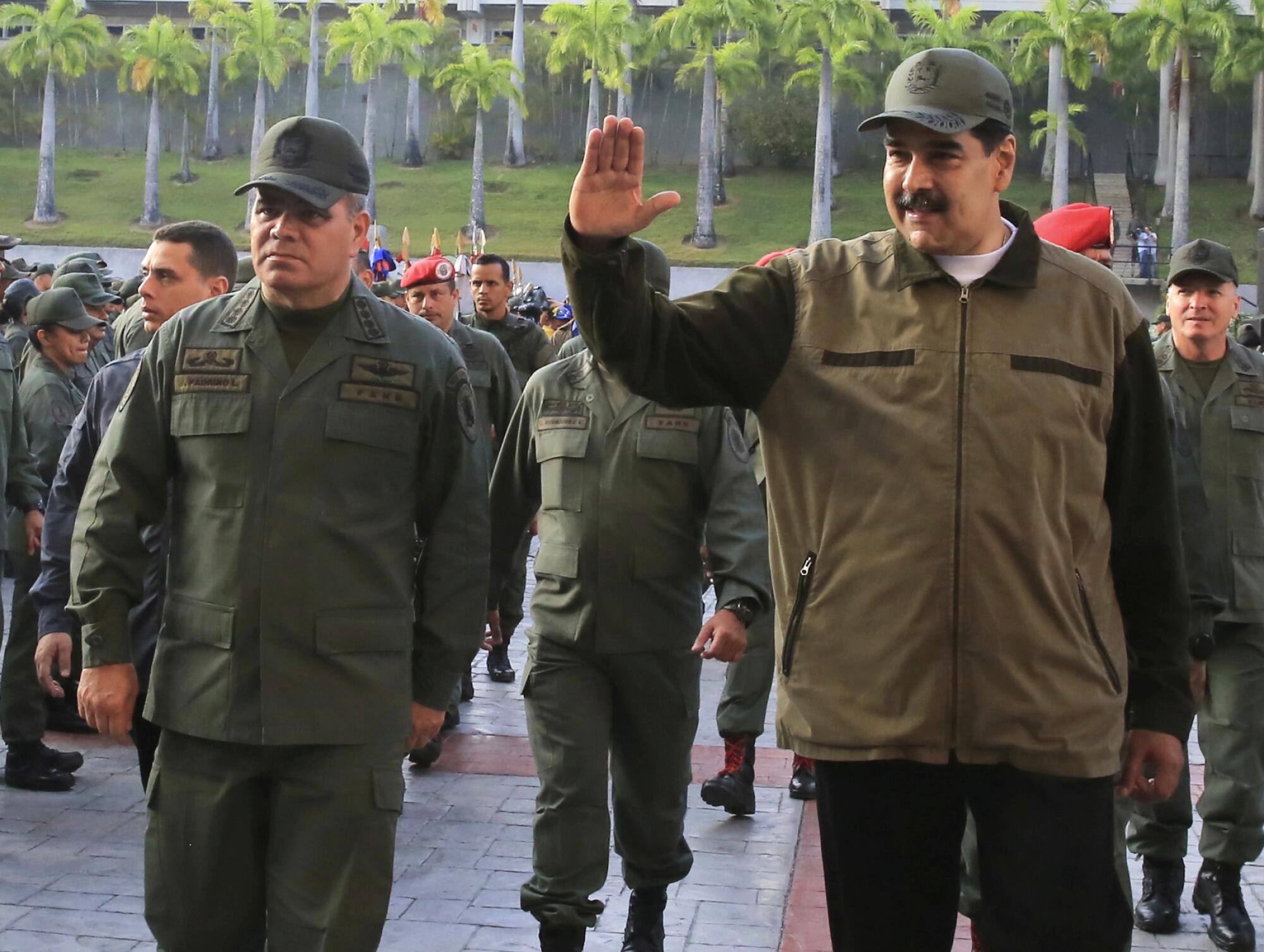 A man with a mustache in khaki-colored jacket and cap waves as other people in military fatigues walk near him 