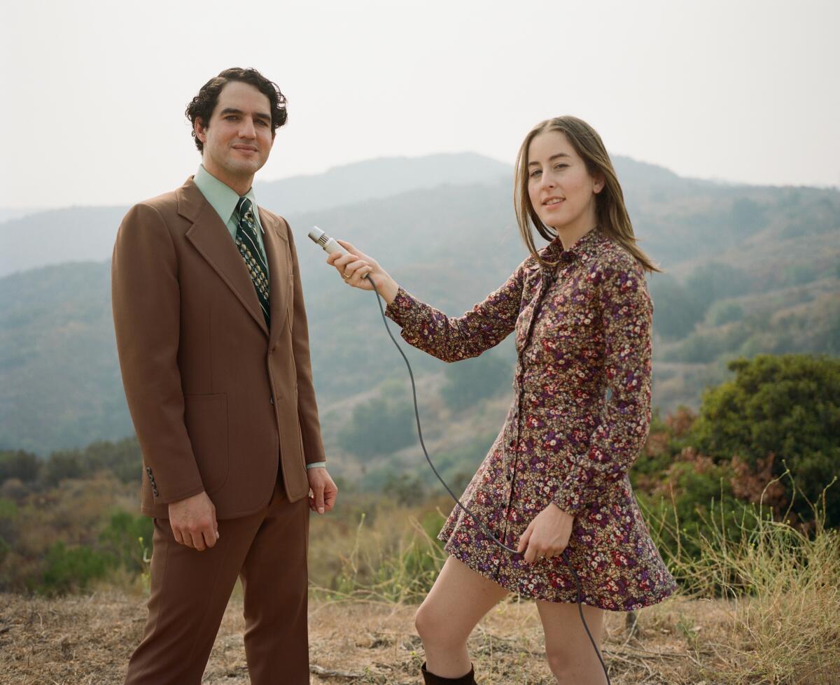 A young woman holds a microphone toward a young man in a suit as they stand on a hilltop