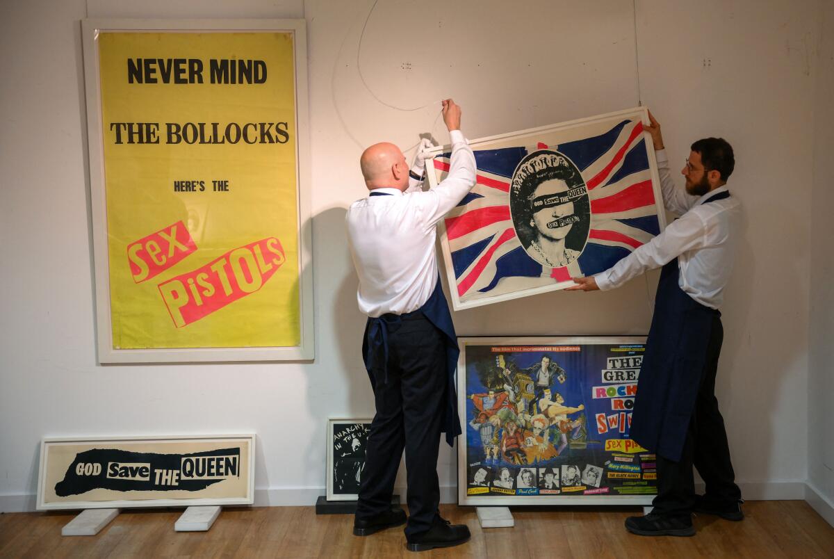 Two gallery assistants are shown hanging a painting that shows Queen Elizabeth's face covered in collaged newsprint