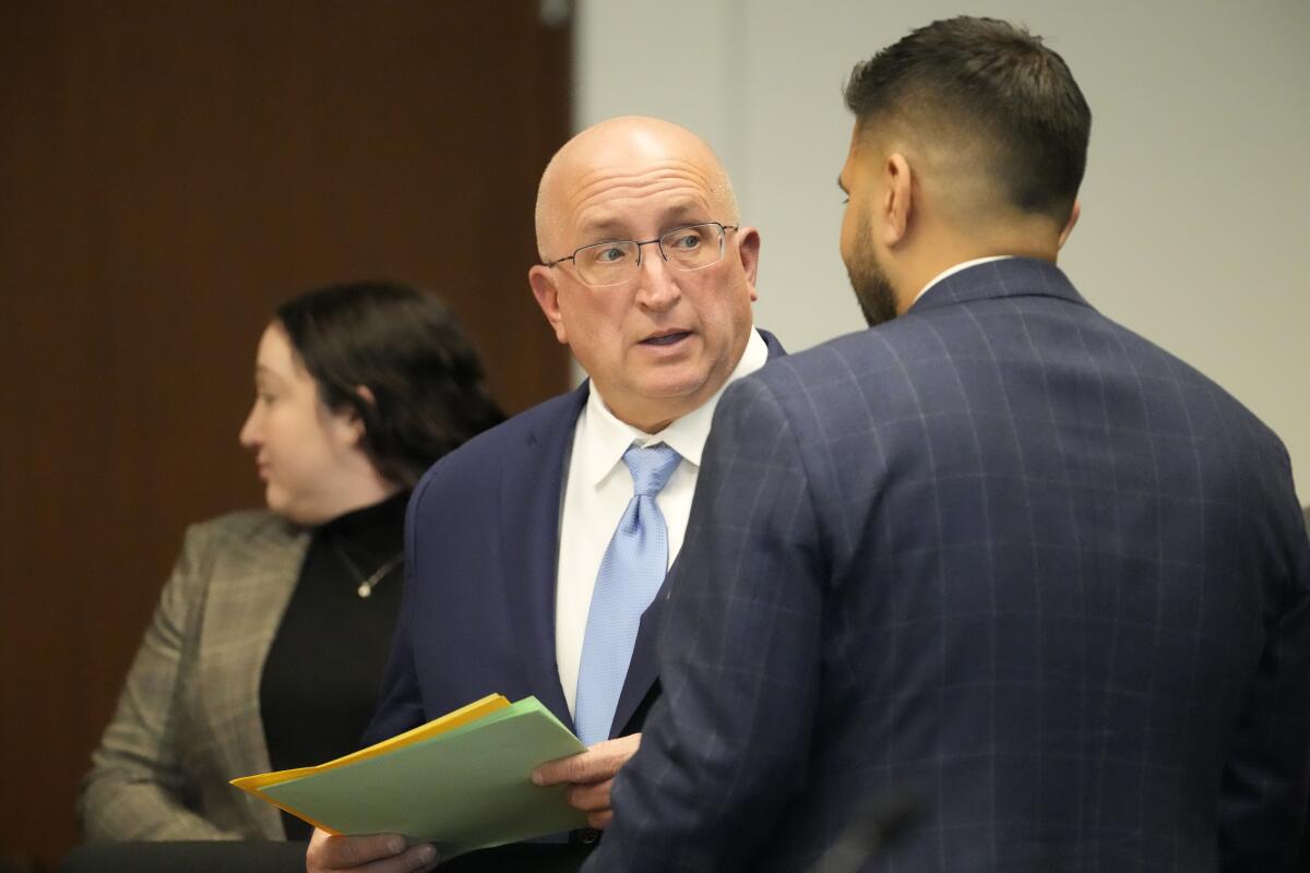 A man in a business suit holding folders talks to another man in a courtroom