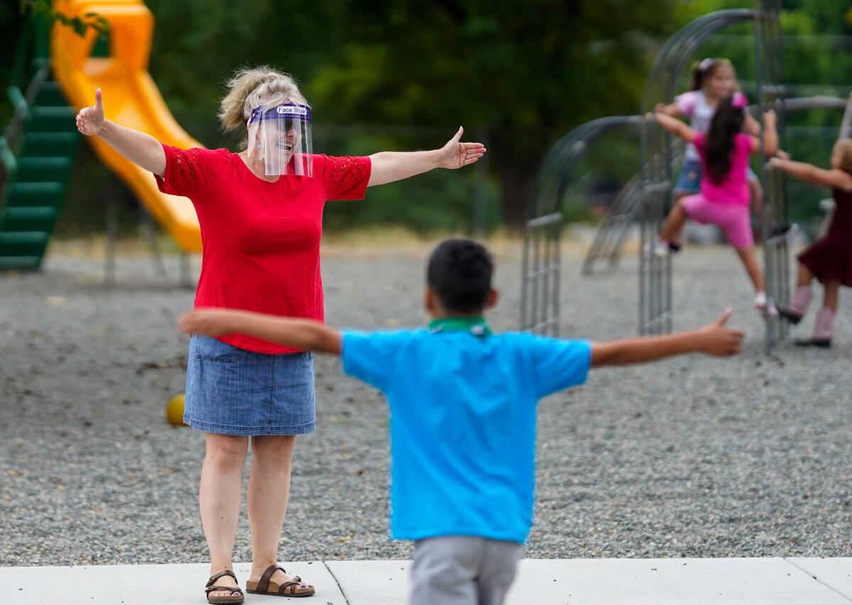 A teacher in a face shield and a student stand several feet apart in a playground, arms stretched wide.