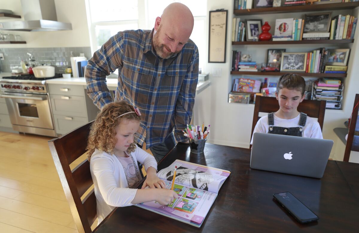 Roy Jensen watches his daughter, Nina, 5, work on a pre-kindergarten activity book, as her sister, Etta, 10, works on an online lesson with some of her fifth-grade classmates though FaceTime, while in their home on Thursday, March 19, 2020 in San Diego.