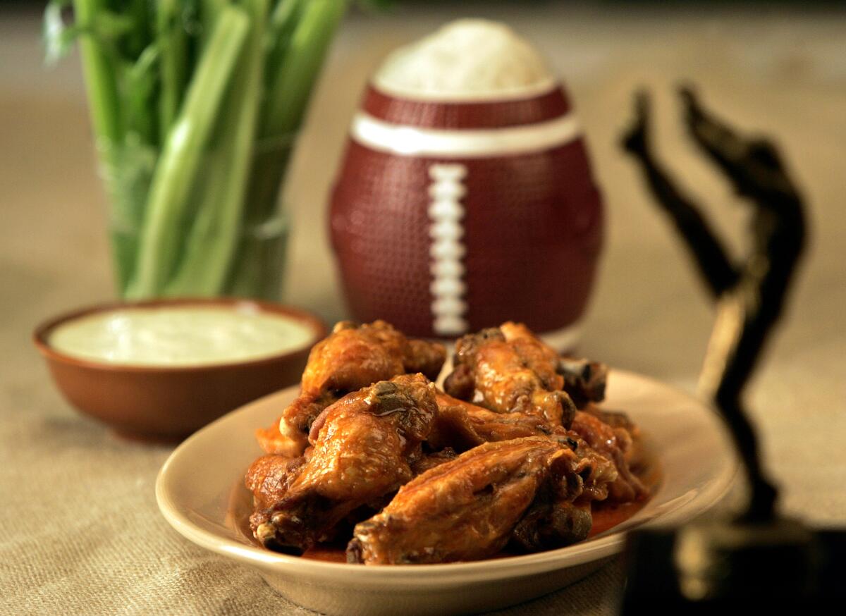 A plate of chicken wings next to a bowl of white sauce, a container of upright celery sticks and a football-shaped mug