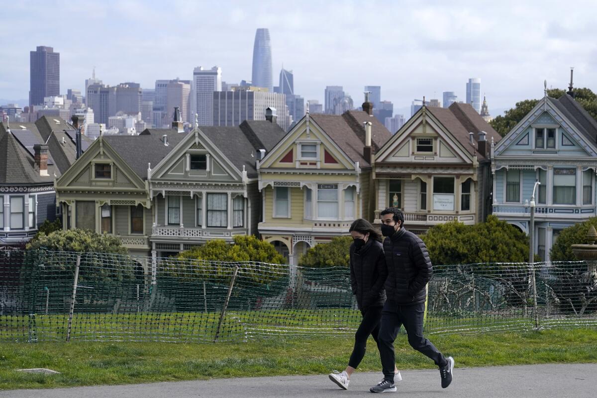 People wear masks while walking in front of a row of Victorian homes.