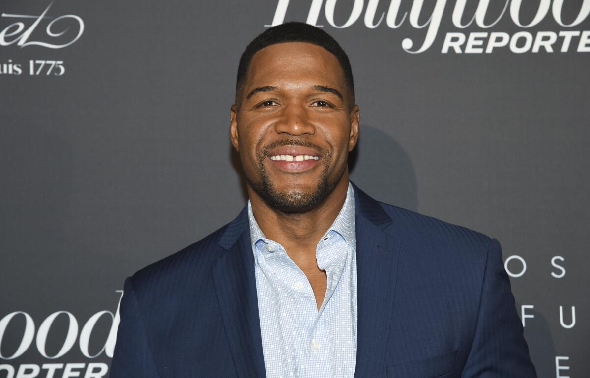 Michael Strahan wears a blue blazer and a baby-blue collared shirt as he stands in front of a gray background