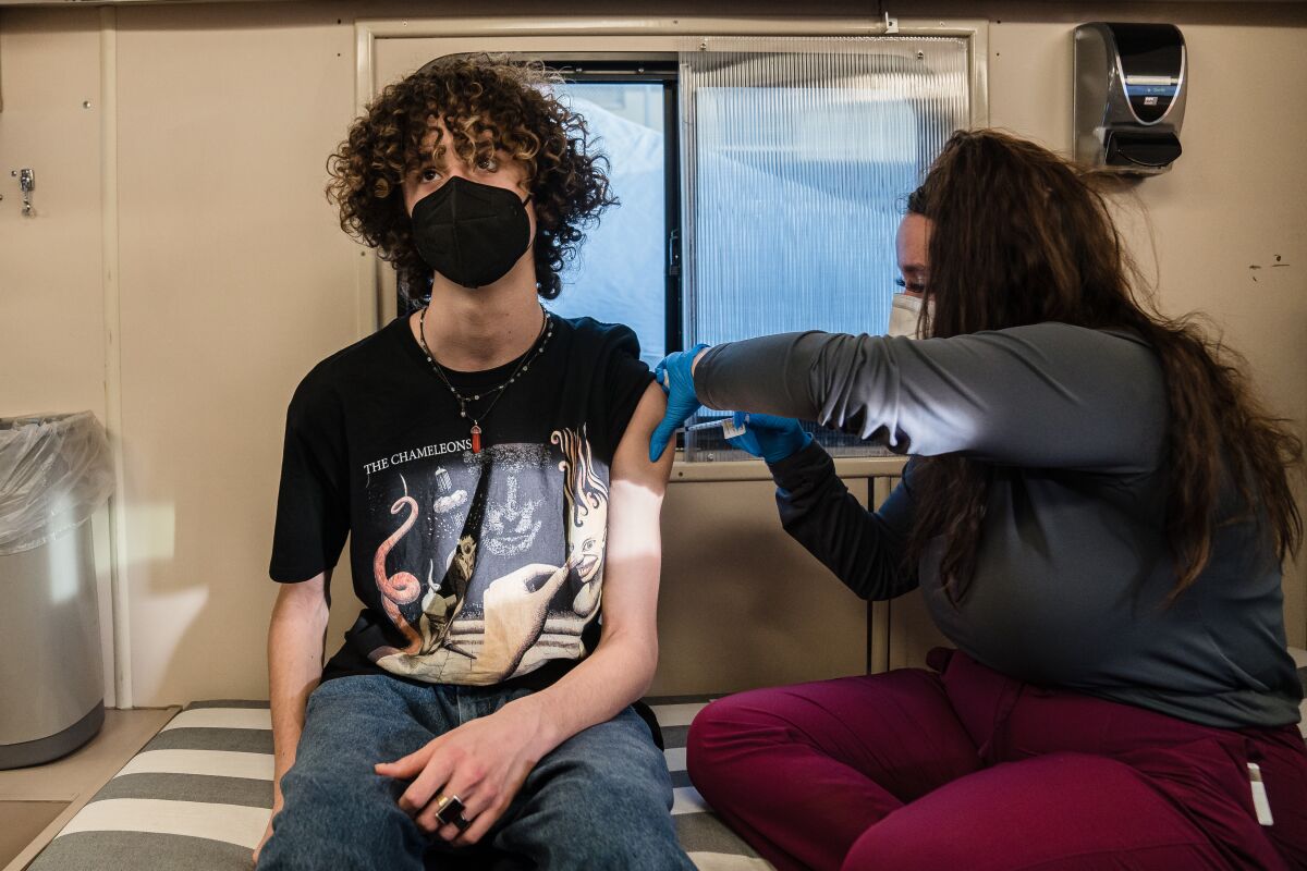 A woman wearing a mask and latex gloves administers a shot to the arm of a teen wearing a mask.