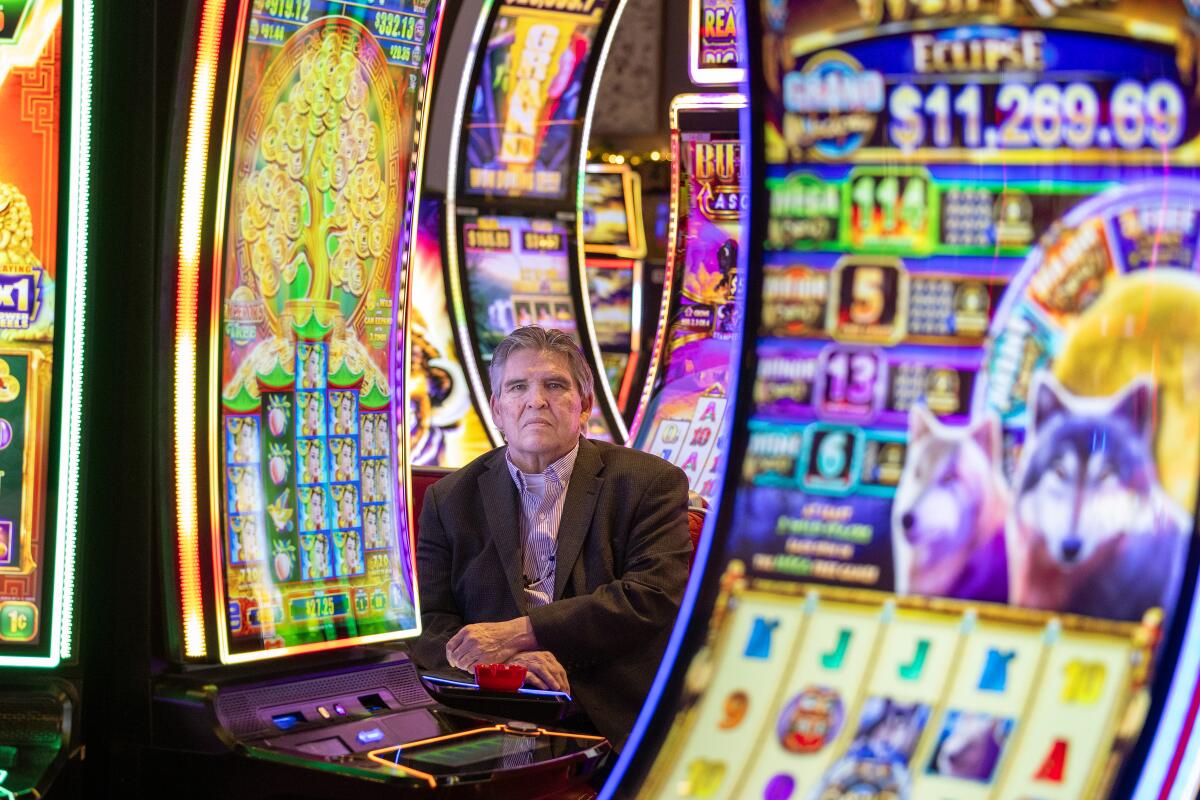 A man sits among slot machines staring at the camera. 