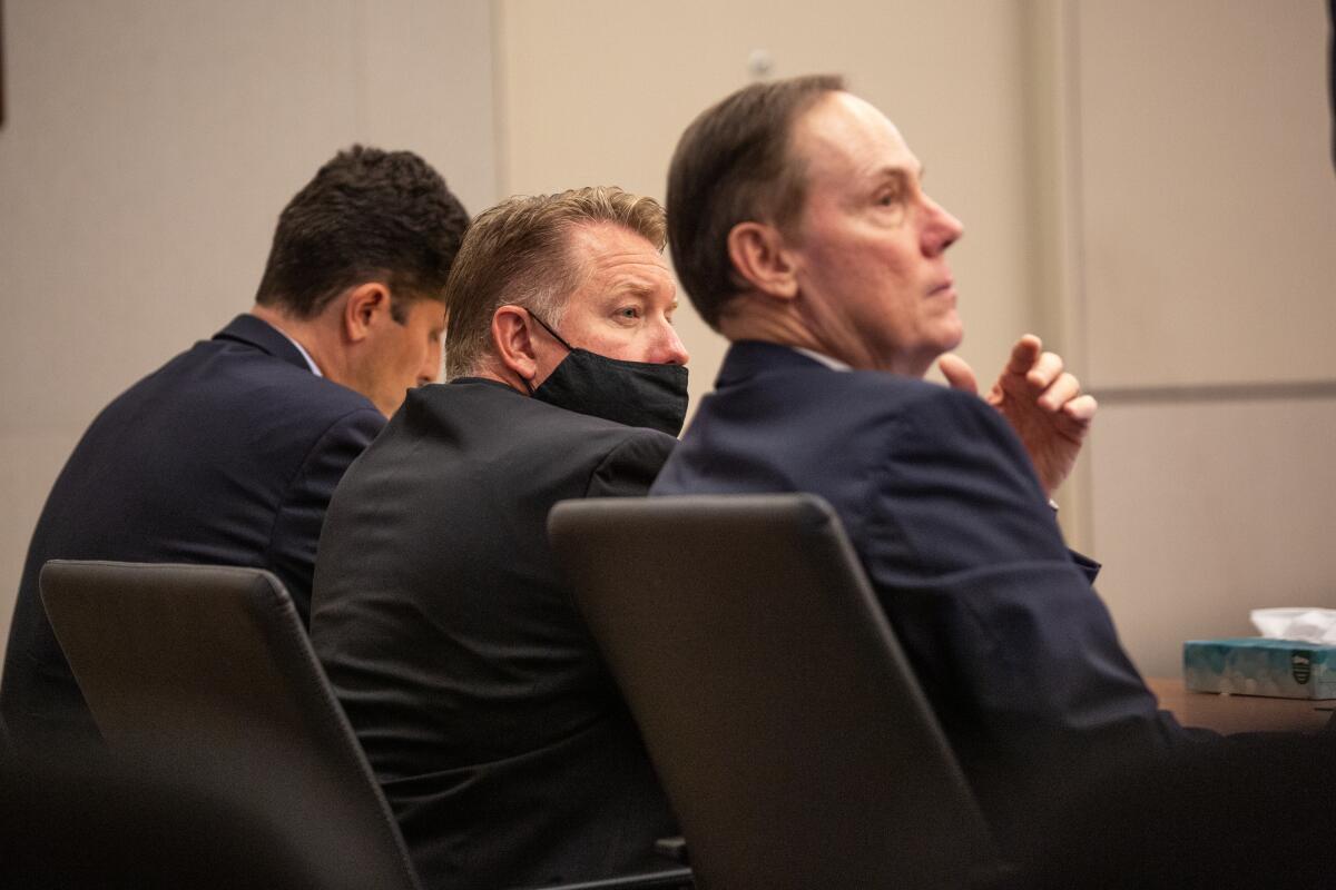 Three men wearing suits sit in a row at a table in a courtroom.