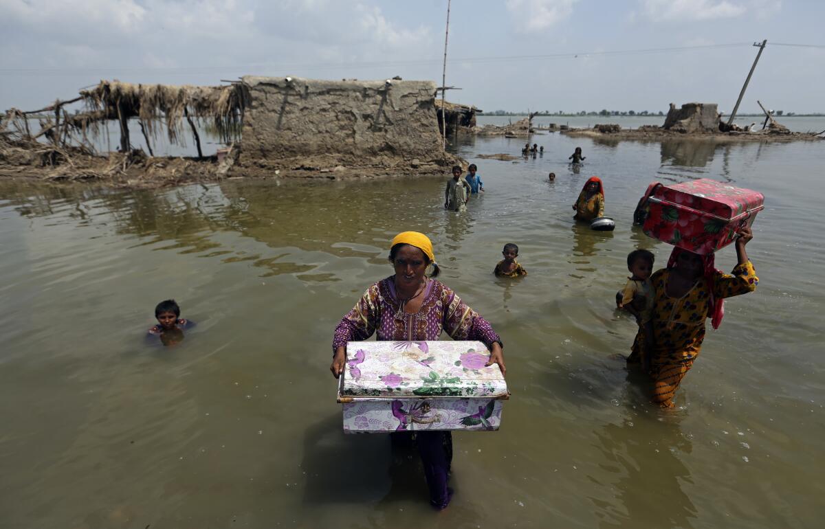 Women carry colorful rectangular containers as they wade in muddy water near flooded homes with thatch roofs