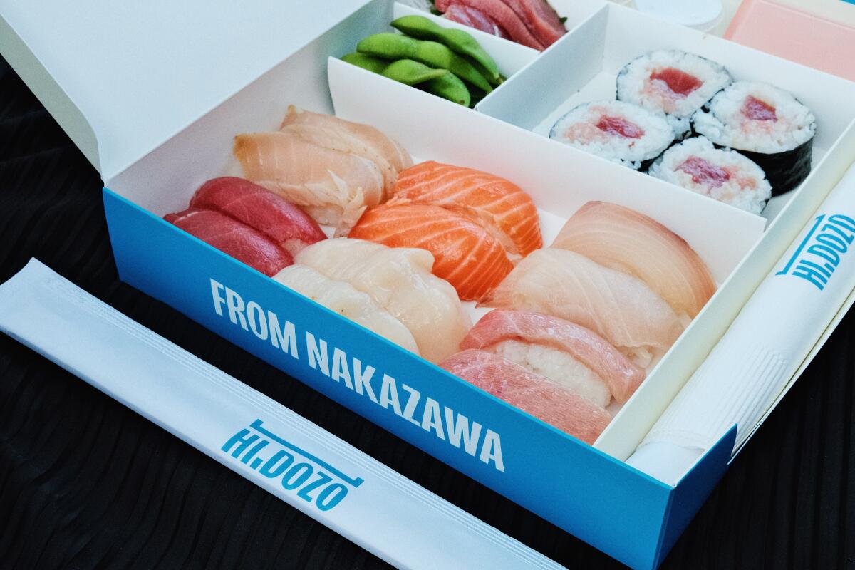 Una fotografía en ángulo de una caja de sushi de papel blanco y azul con 12 piezas de nigiri, cuatro de maki y sashimi con edamame.