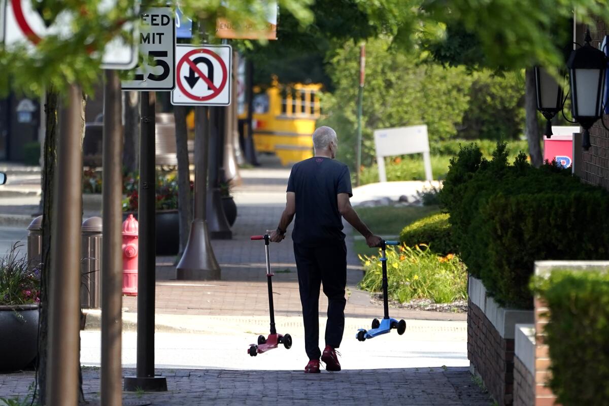 A man walks down a sidewalk carrying two small scooters.