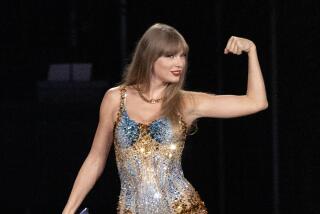 Taylor Swift in a bejeweled blue and gold leotard flexing her left arm and holding a microphone in her right hand on a stage
