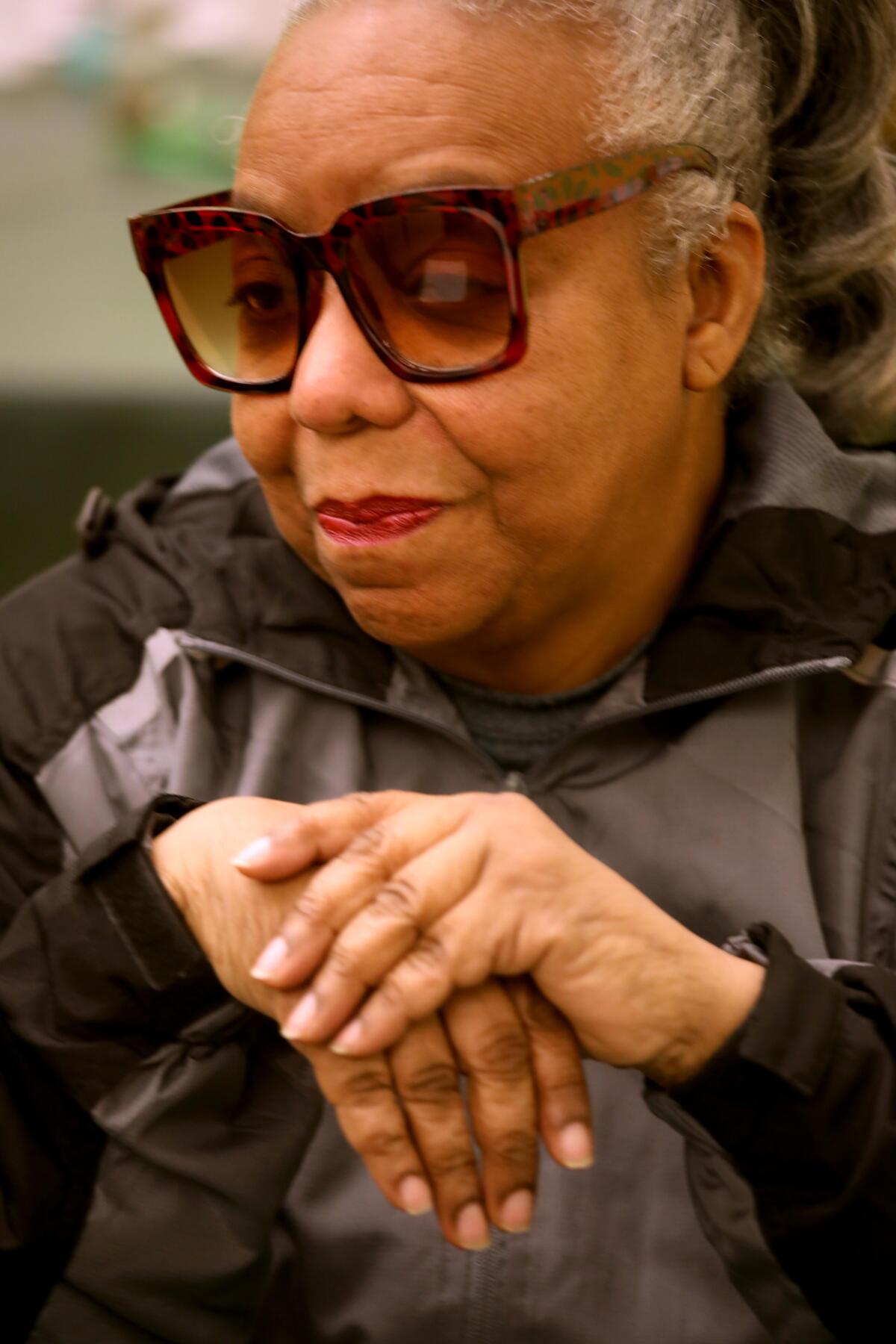 A woman with large sunglasses sits with her hands clasped in front of her.