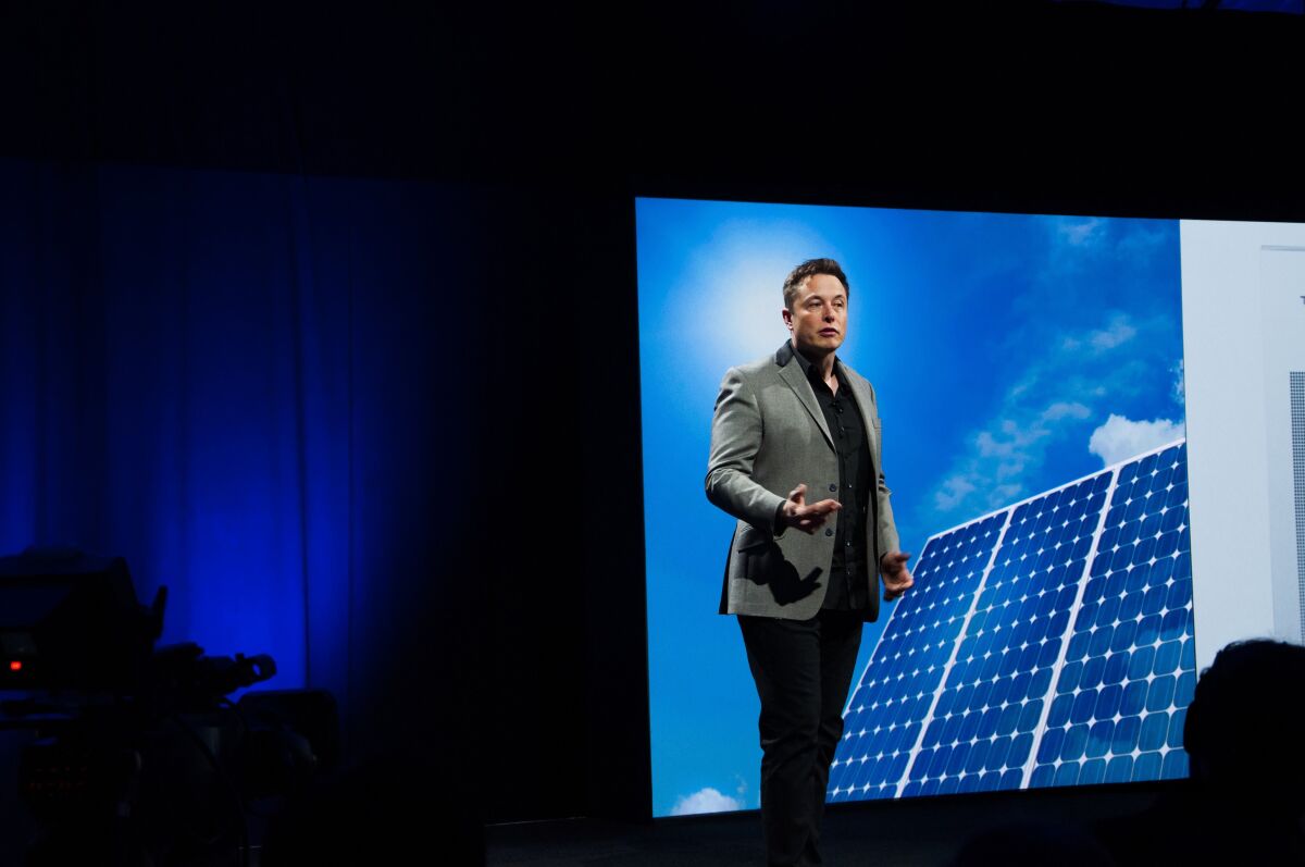 Elon Musk speaks in front of an image of solar panels