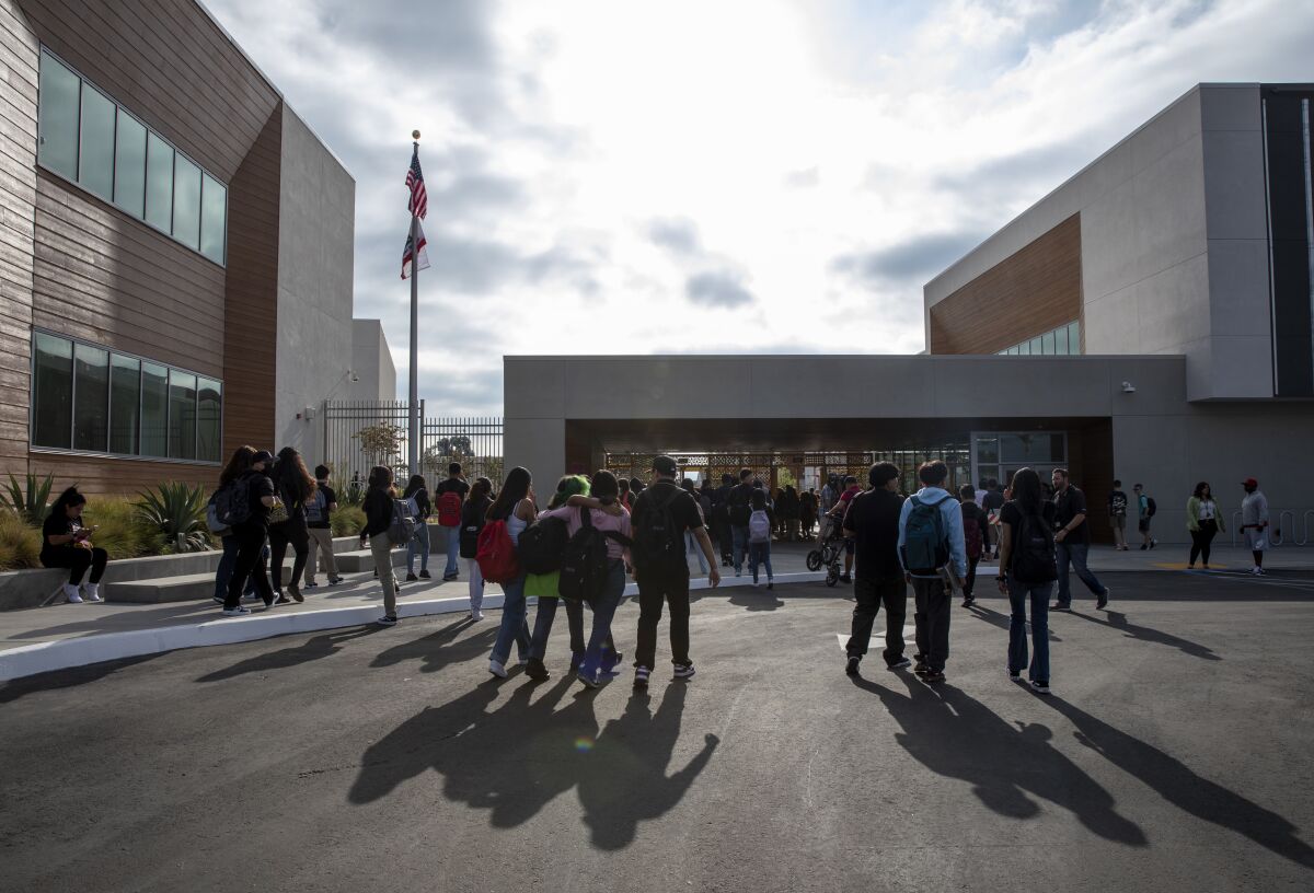 Dozens of teen students wearing backpacks stream toward a school building against a cloudy morning sky.
