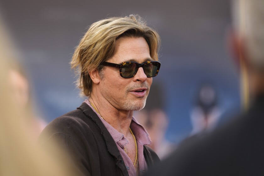A blond man in sunglasses and a pink shirt under a brown jacket among people.