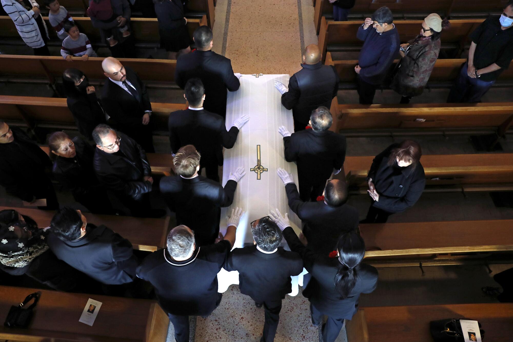 A view from above of people in dark suits flanking a white coffin occupying the aisle between pews with people standing