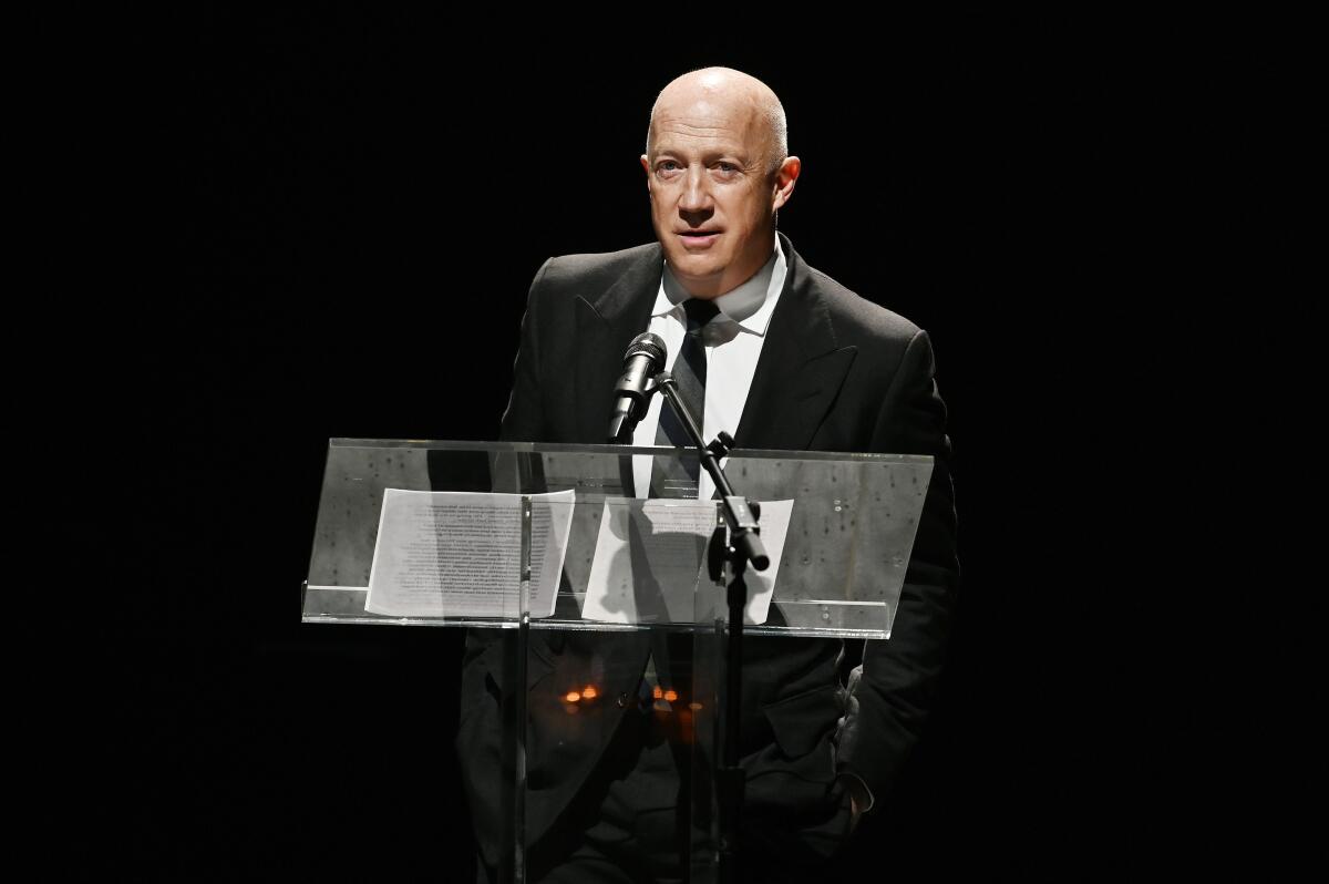 A man in a suit and tie speaks at a lectern on a dark stage 