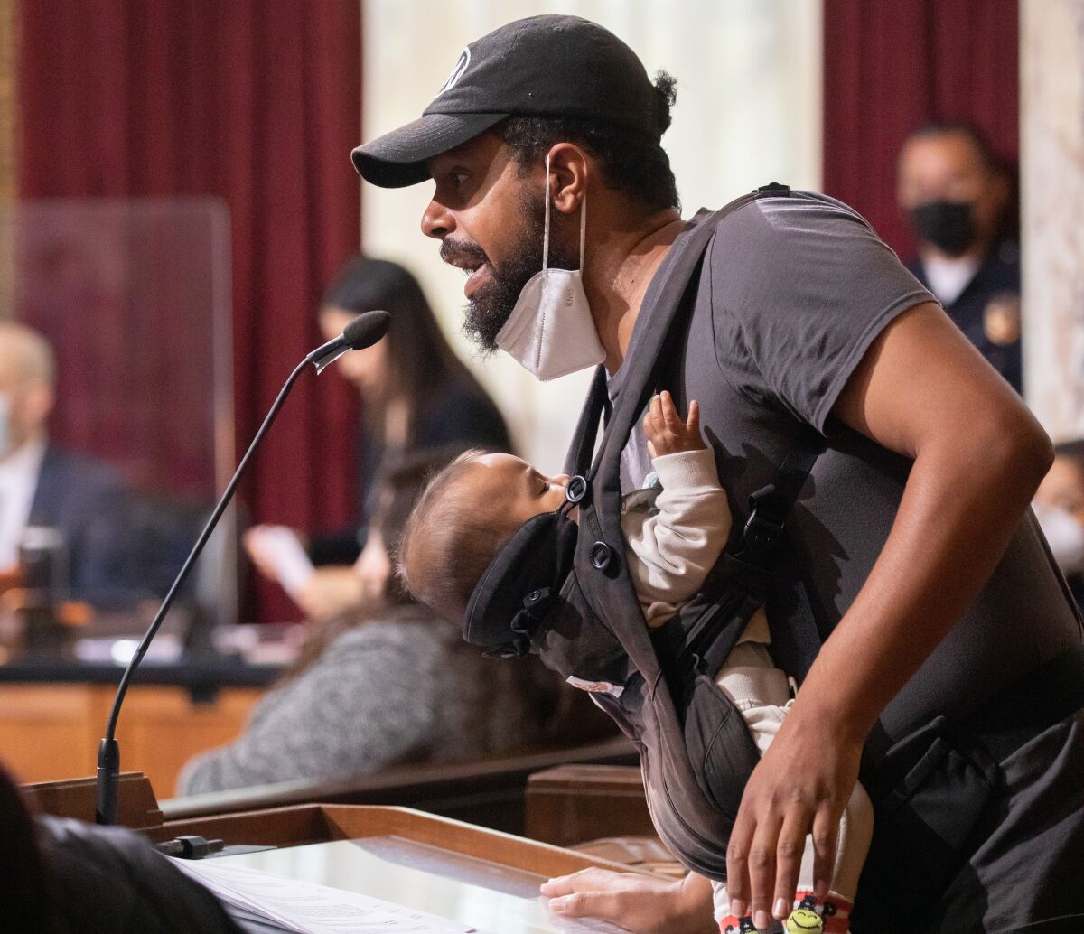 A man with a baby in a sling on his front speaks into a microphone on a lectern