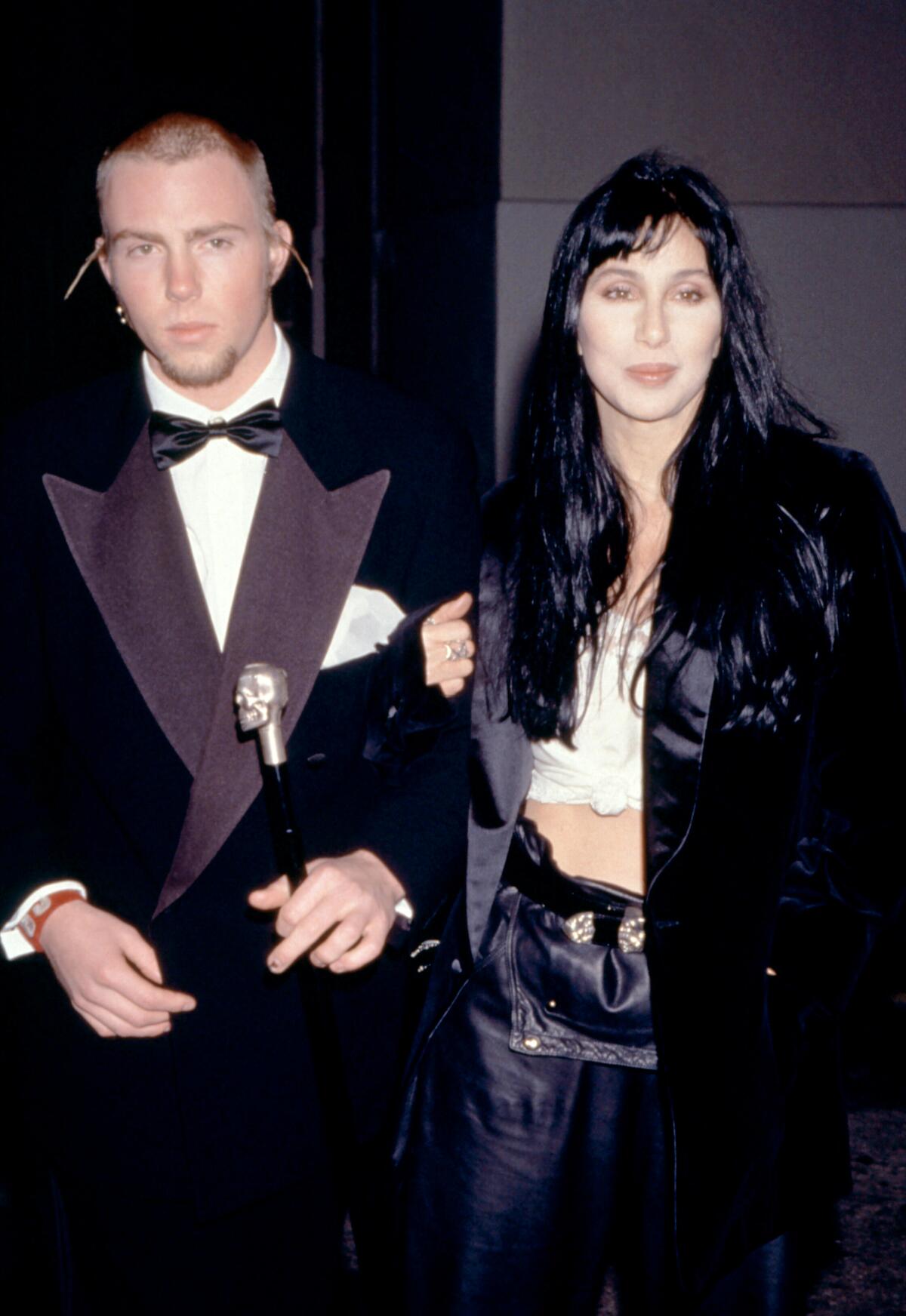 Elijah Blue Allman wears a tuxedo and links arms with his mother Cher, wearing a long black coat and white crop top
