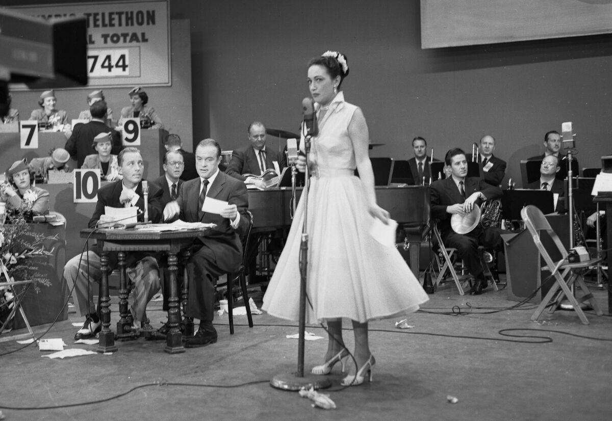 A woman speaks at a microphone and two men sit at a table; behind them are a band and people answering phones.