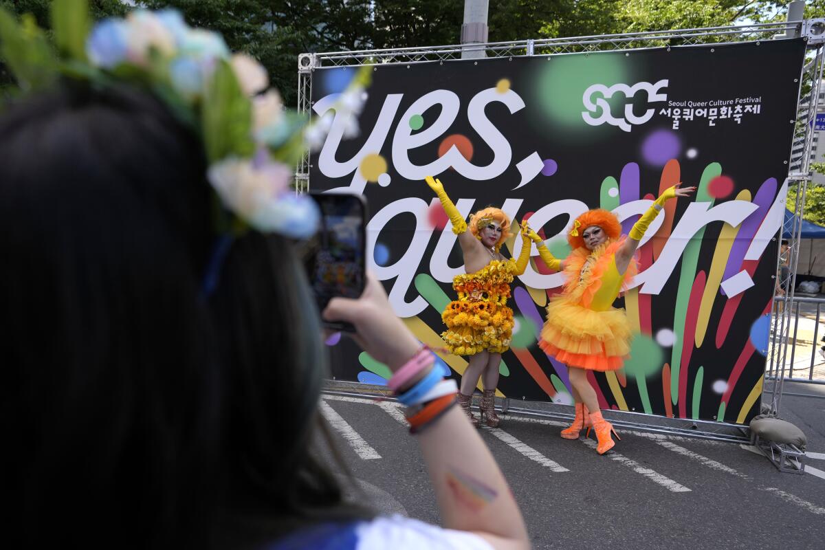Participants in elaborate yellow and orange outfits pose for a souvenir photo in front of a sign that says yes, queer.