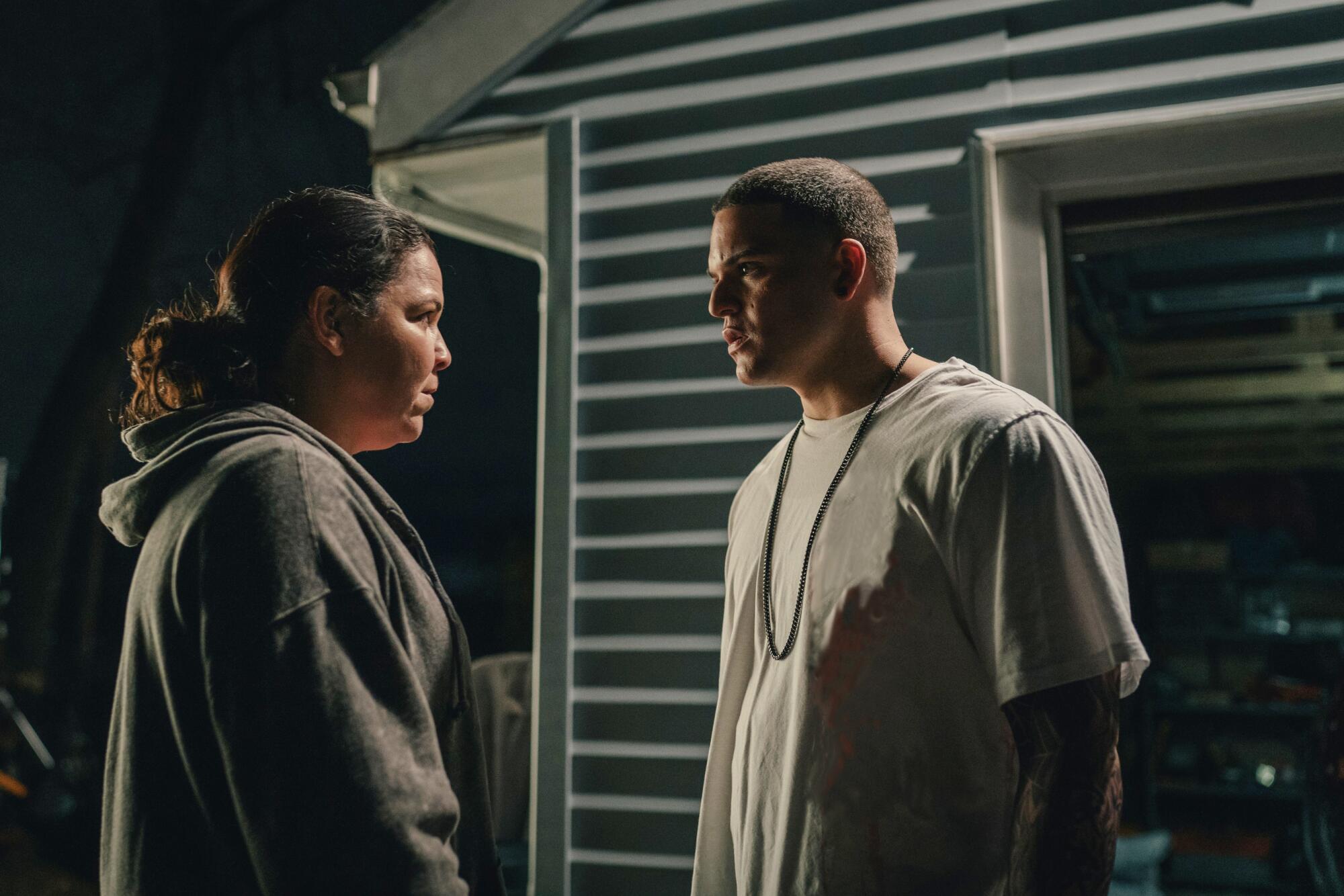 A woman in a ponytail and hoodie looks at a man wearing a chain and white shirt.