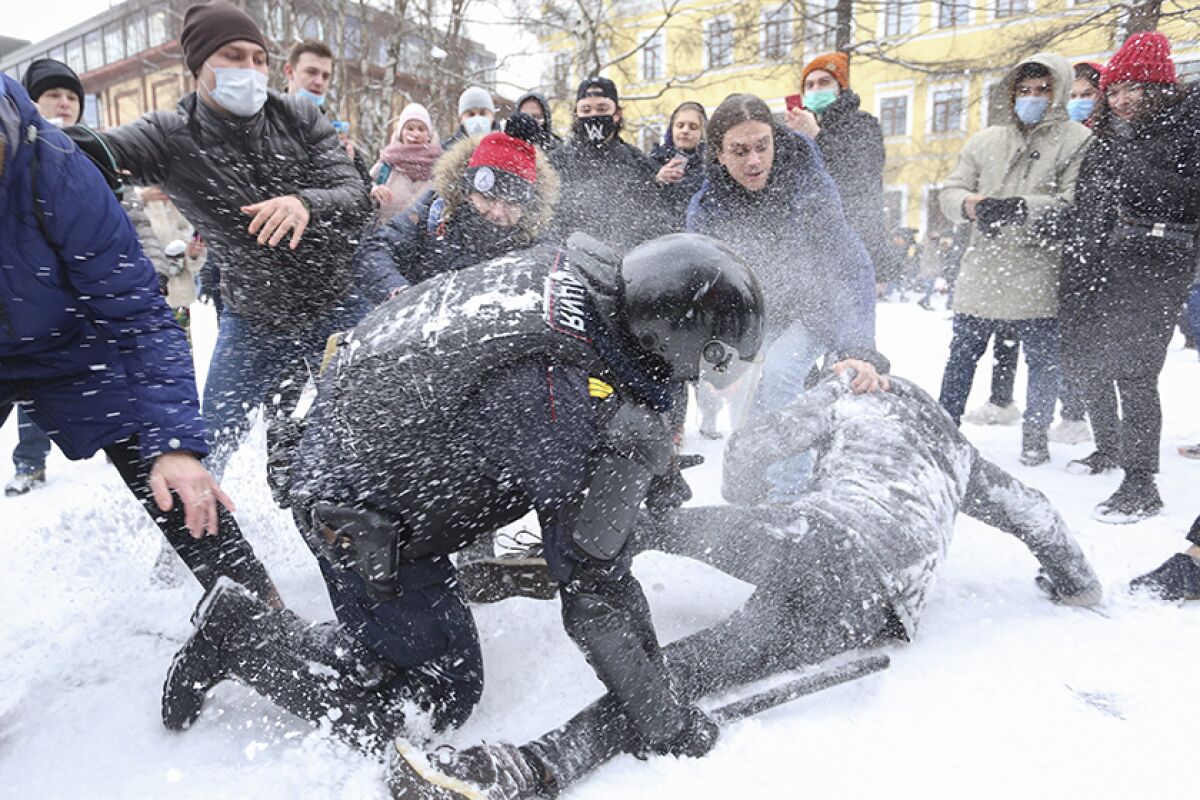 Amid a crowd and snow, a policeman with a baton leans on one knee over a man.
