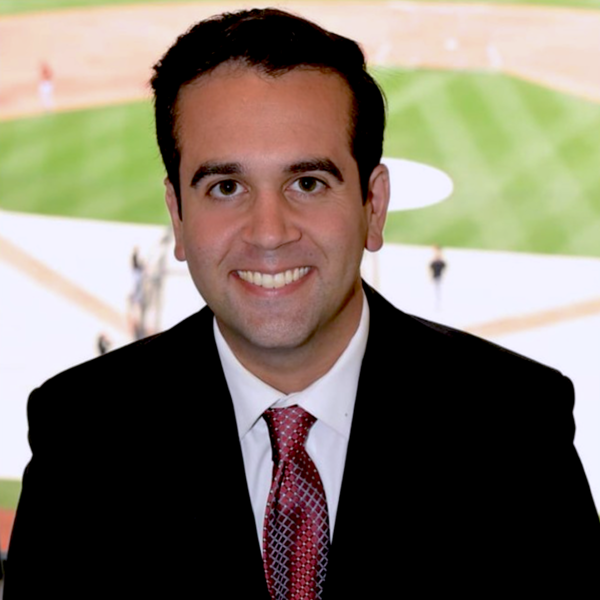 Man in a suit and tie smiles as he sits in a television booth above a baseball field 