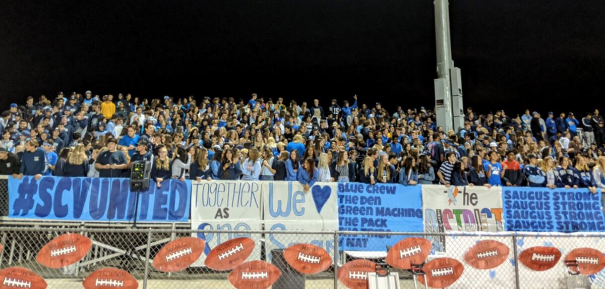 A crowd of fans from the Santa Clarita Valley at a West Ranch high school football game holding messages including "#SCVUnited" and "Saugus Strong."