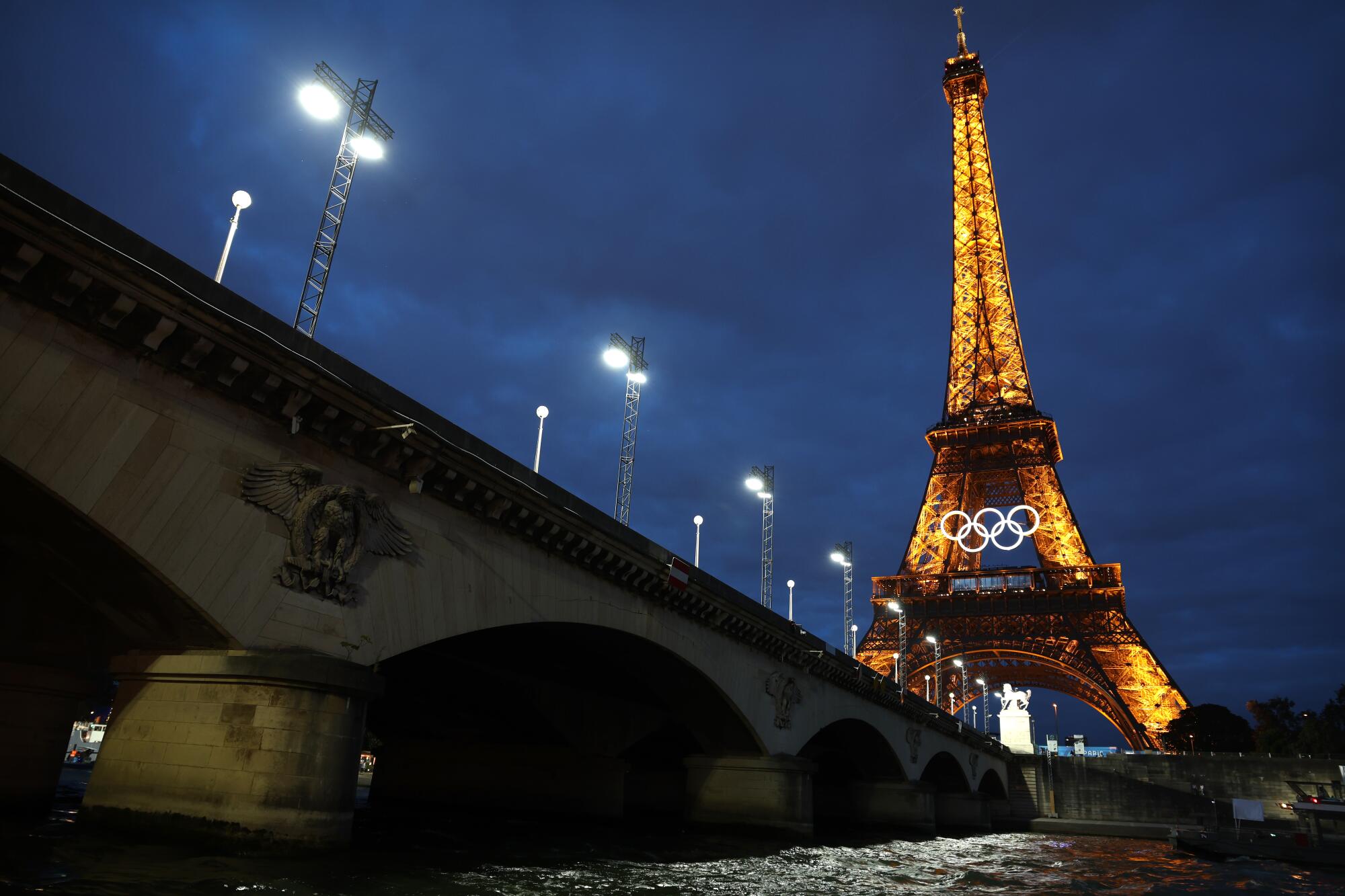 The Eiffel Tower shines brightly at night as the Seine River rushes under a bridge nearby.