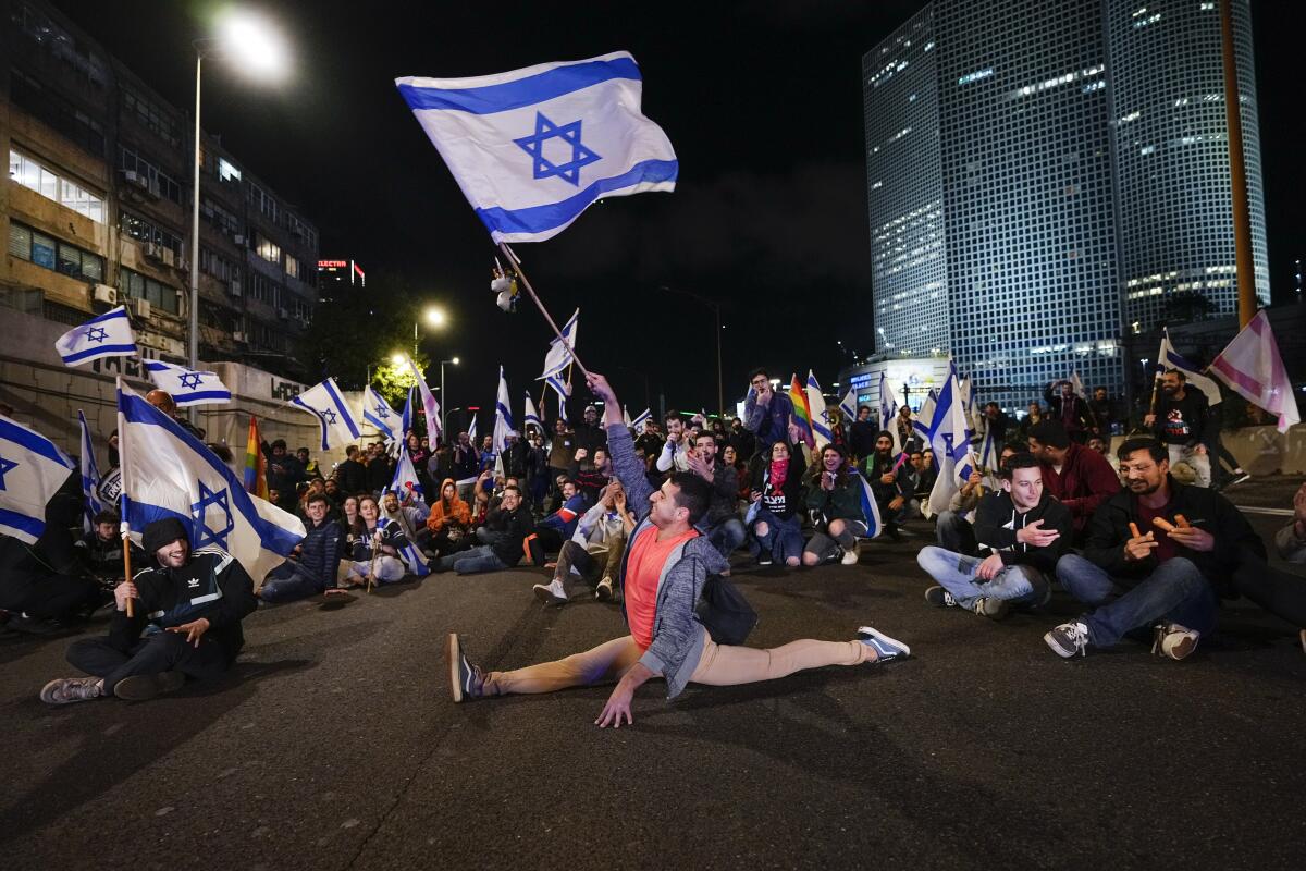 A man holding up an Israeli flag does the splits on a highway in Tel Aviv as others sit during a protest 