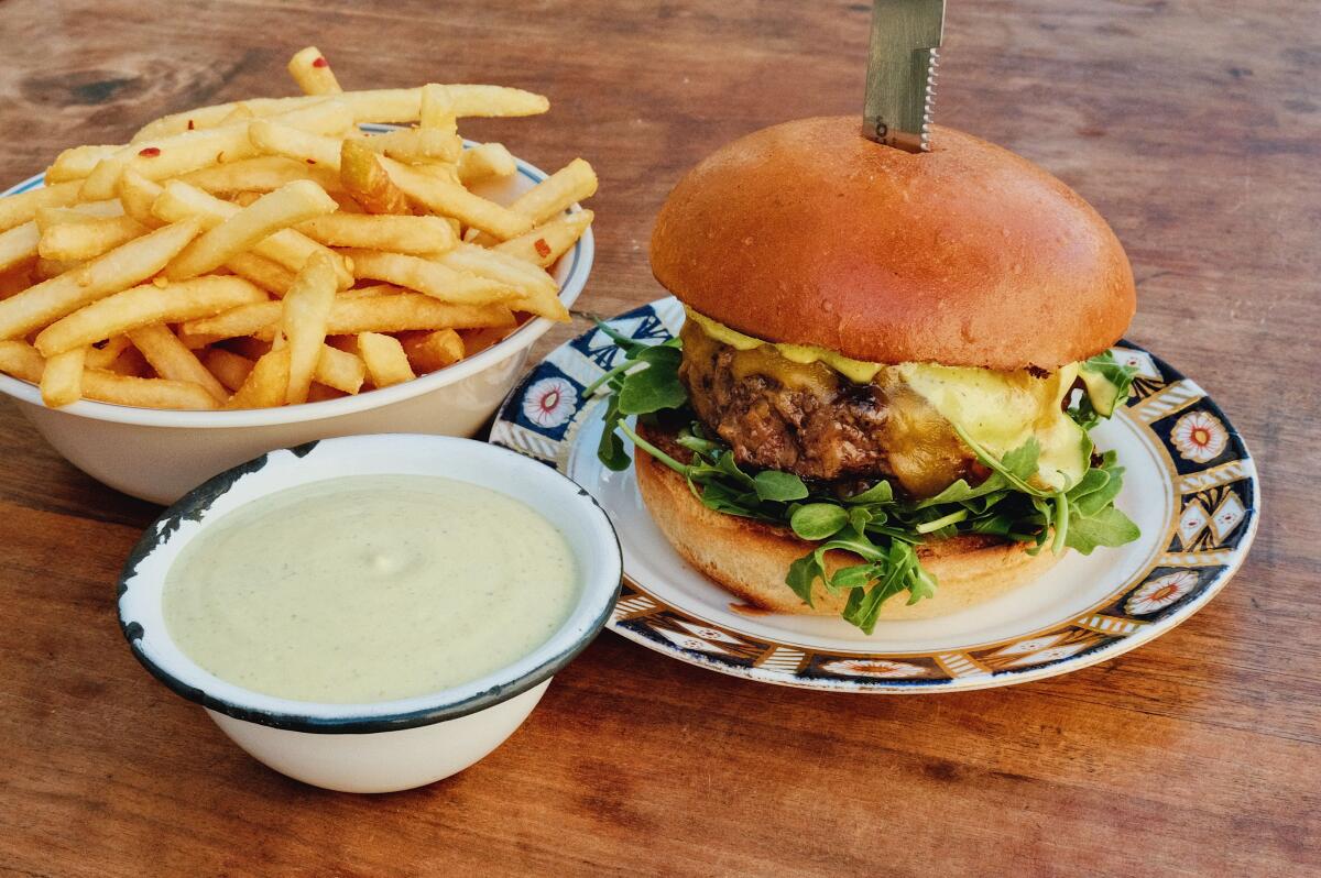A knife sticks out of a thick cheeseburger's top bun, on a wood table next to separate dishes of fries and off-white sauce