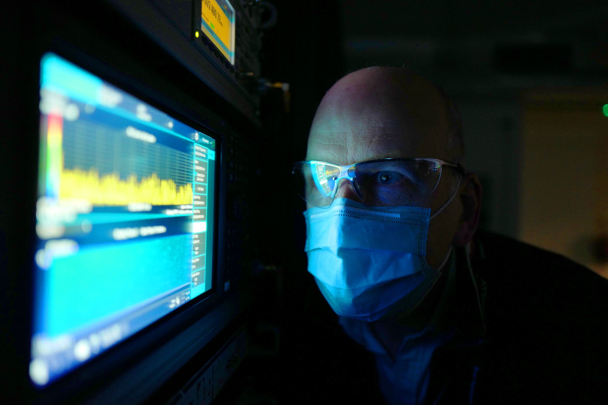 A man in glasses and a mask is illuminated by glow of a screen