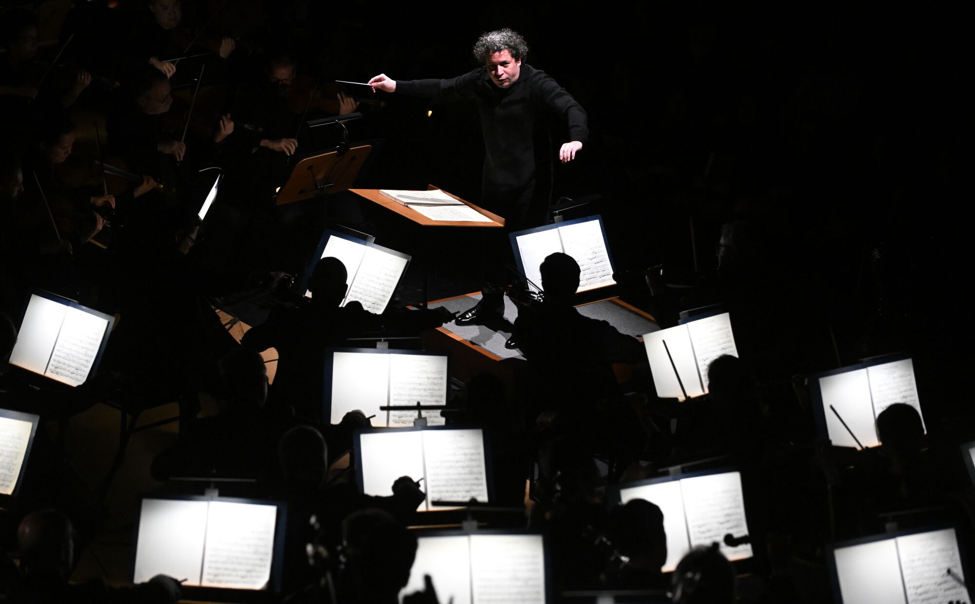 Seen in upper part of the frame against black background, Gustavo Dudamel directs the orchestra below.