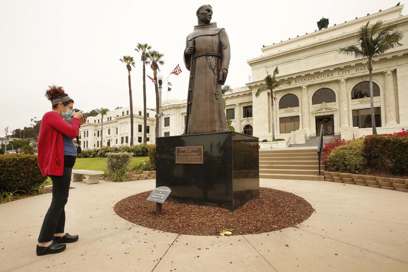 VENTURA, CA - JUNE 24: Erica Cohee from Thousand Oaks photographs the bronze statue of Father Junipero Serra who founded nine Spanish missions in California including Mission San Buenaventura as it stands in front of Ventura City Hall. A joint statement was issued from local political, indigenous and Catholic leaders pledging to remove the monument, originally commissioned in the 1930s but replaced in 1989. "The time has come for the statue to be taken down and moved to a more appropriate nonpublic location," read the statement, issued by Ventura Mayor Matt LaVere. After a protest last weekend Ventura city spokeswoman Heather Sumagaysay said the city is making plans to host community discussions regarding the Serra statue and there is no timeline for its removal, she said. "It is our priority to be receptive to public concerns and provide an environment where all voices are heard and respected. A historic decision such as this will involve the voices of the Chumash tribe, the City Council and the residents of Ventura," Sumagaysay said in an emailed statement. "The city remains committed to collaborating with the community to determine next steps. We will inform the public of opportunities to participate and offer input at a future meeting." The move comes after a Father Serra statue was toppled last weekend on Olvera St. in downtown Los Angeles and people were considering changing the name of Ft Bragg, but voted the change down. Ventura City Hall on Wednesday, June 24, 2020 in Ventura, CA. (Al Seib / Los Angeles Times)
