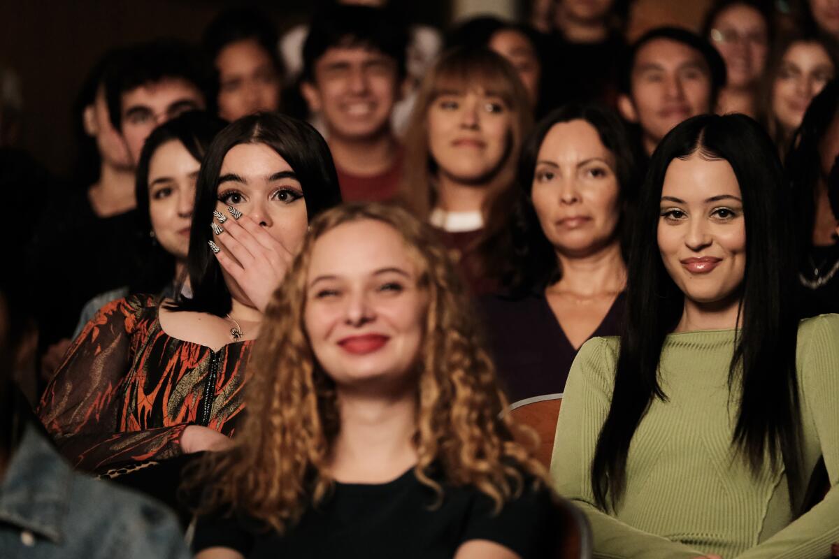 Barbie Ferreira covers her mouth with her hand and Alexa Demie smirks while seated among a crowd of people.
