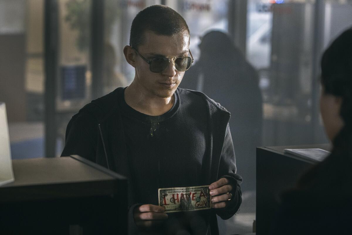 In "Cherry," Tom Holland, wearing dark sunglasses, shows a bank teller a dollar bill on which "I have a gun" is written.