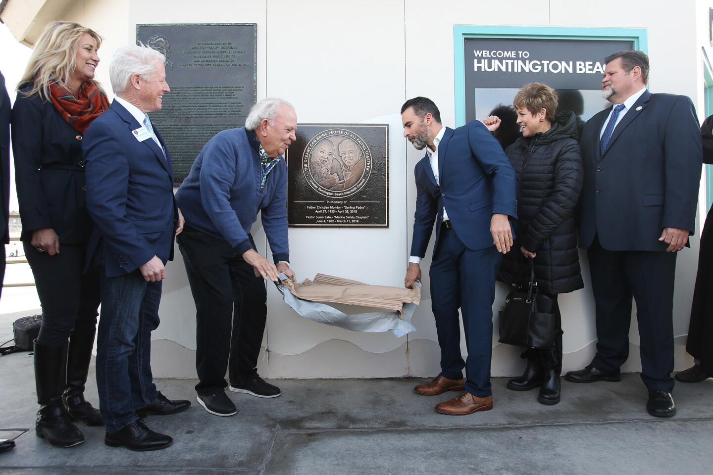 A plaque honoring late Huntington Beach pastors and surfing icons Christian Mondor and Blaine "Sumo" Sato is unveiled by Dennis Gallagher and Cesar Gutierrez and members of the City Council during a dedication ceremony Wednesday on the Huntington Beach Pier.
