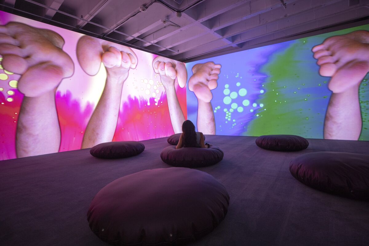 An installation view shows projections of blobby color and human feet occupying the entire corner of a large gallery.