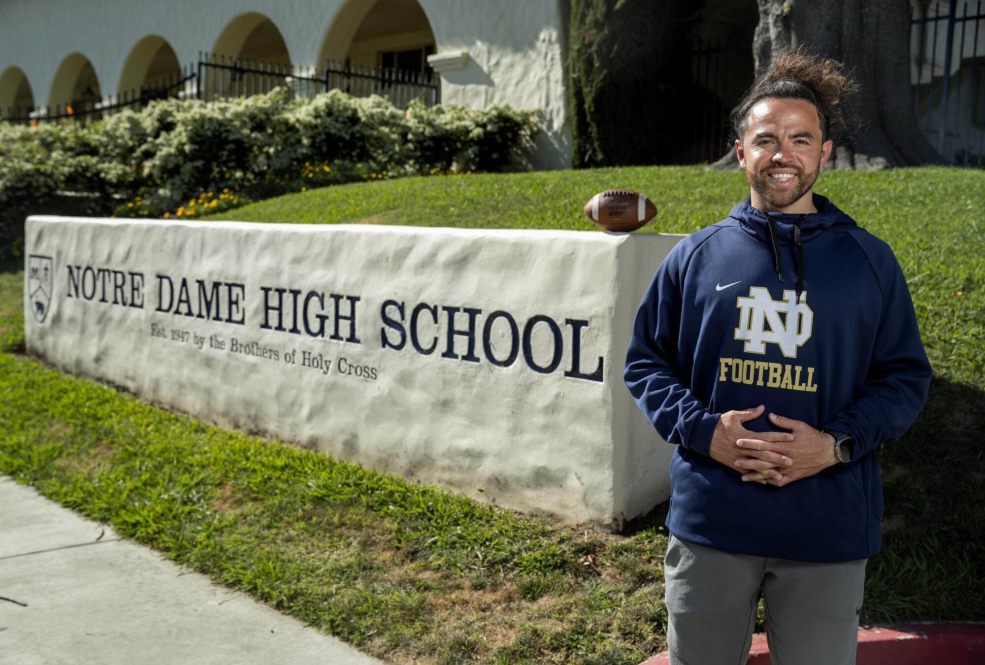 Evan Yabu poses in front of a sign on campus reading "Notre Dame High School" with a football on top
