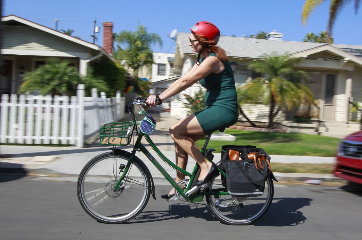 A woman in a green dress and red helmet pedals her e-bike past houses on a residential street