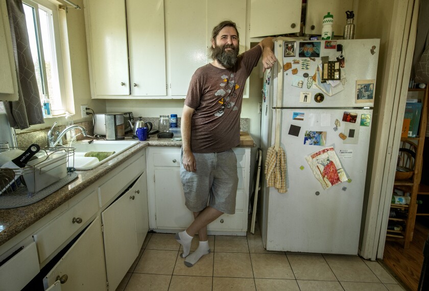 A man in a kitchen leans against a refrigerator and smiles 