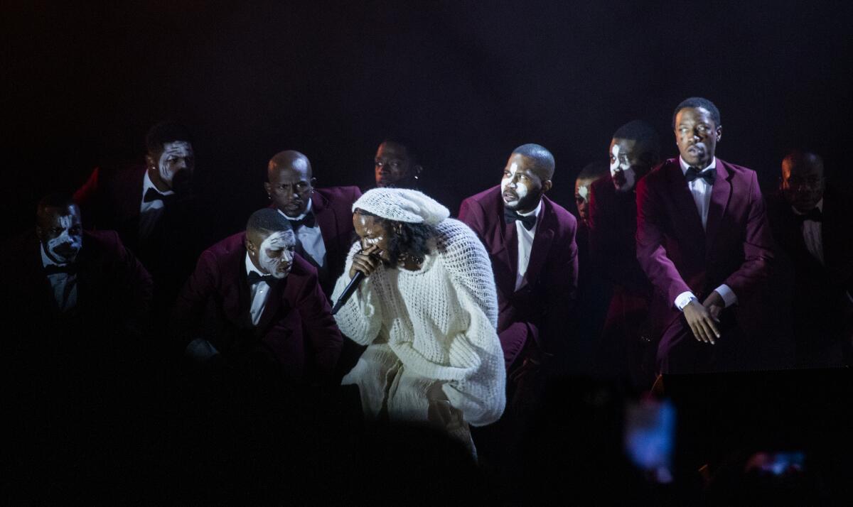 A man, rapping into a microphone, is surrounded by men in dinner jackets and ties, some with white face makeup.