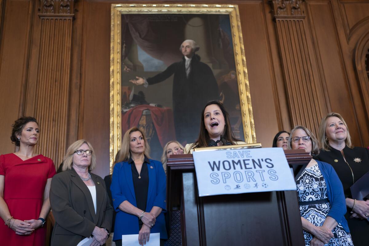 A woman speaks from a lectern with a sign reading "Save women's sports" and several other women behind her.